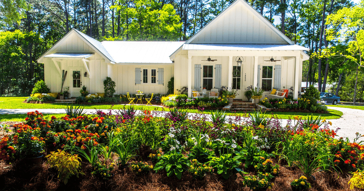 A white house with a front porch surrounded by a well-maintained garden filled with colorful flowers and lush greenery. The scene is set on a sunny day with trees in the background.