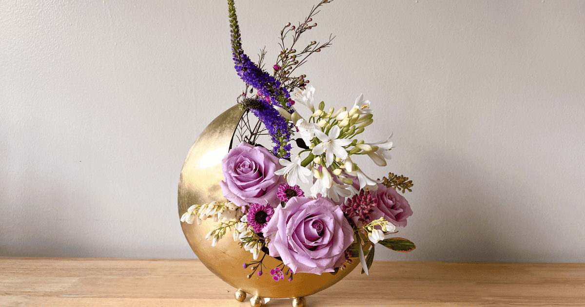A floral arrangement with purple roses, white flowers, and assorted greenery in a round, gold vase on a wooden surface.