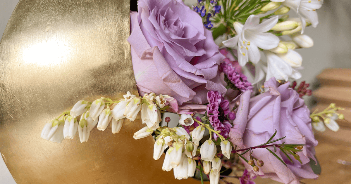 A close-up of a floral arrangement with light purple roses, white flowers, and greenery in a metallic gold vase.