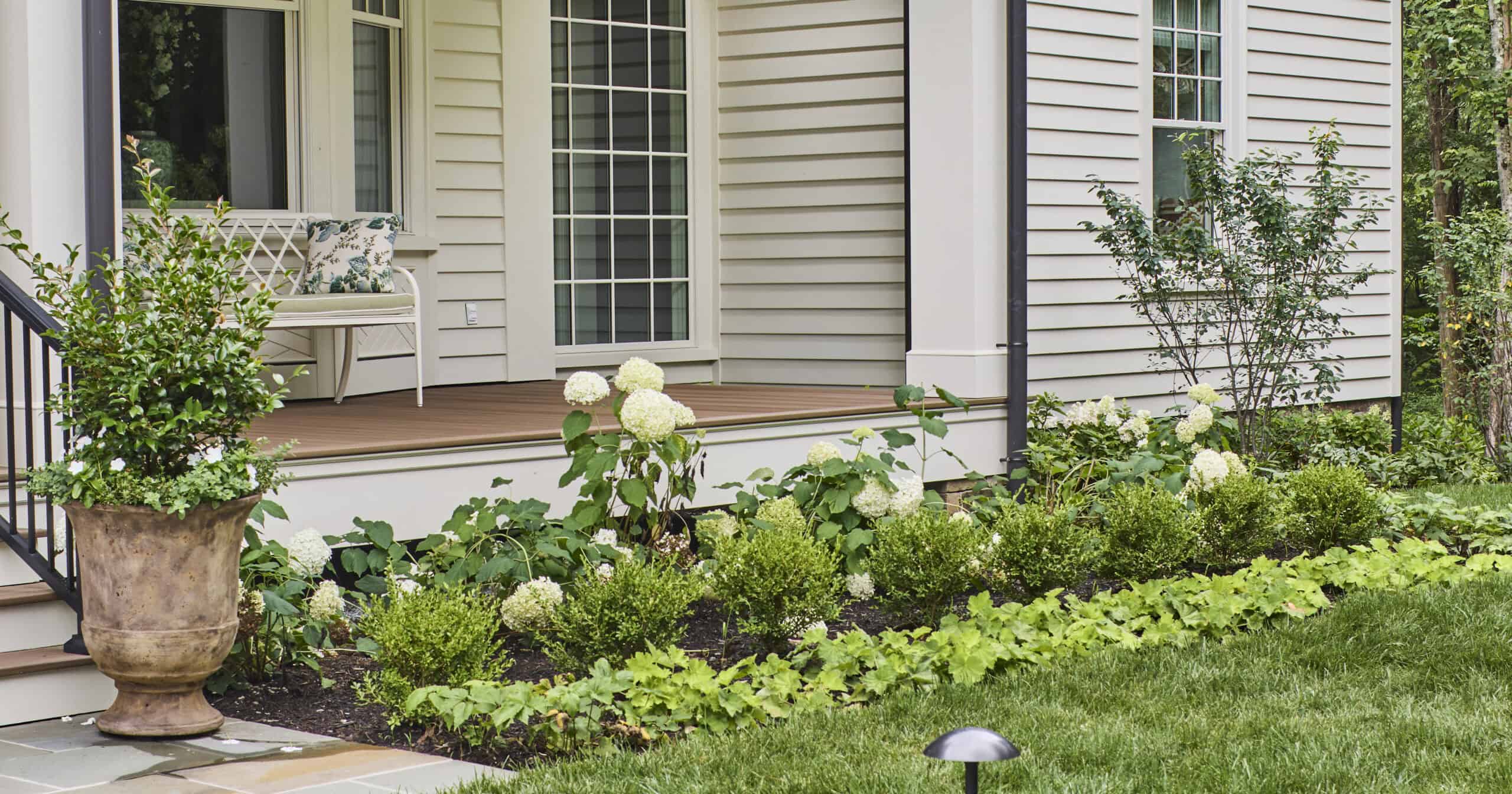 Front entry garden bed with layered foundation plantings, including a row of Better Boxwood – Babylon Beauty™ for structure, behind which White Wedding® and Sweet Starlight™ Hydrangeas bloom with large white flowers. A Diamond Spire® Gardenia is featured in a large planter near the porch stairs.