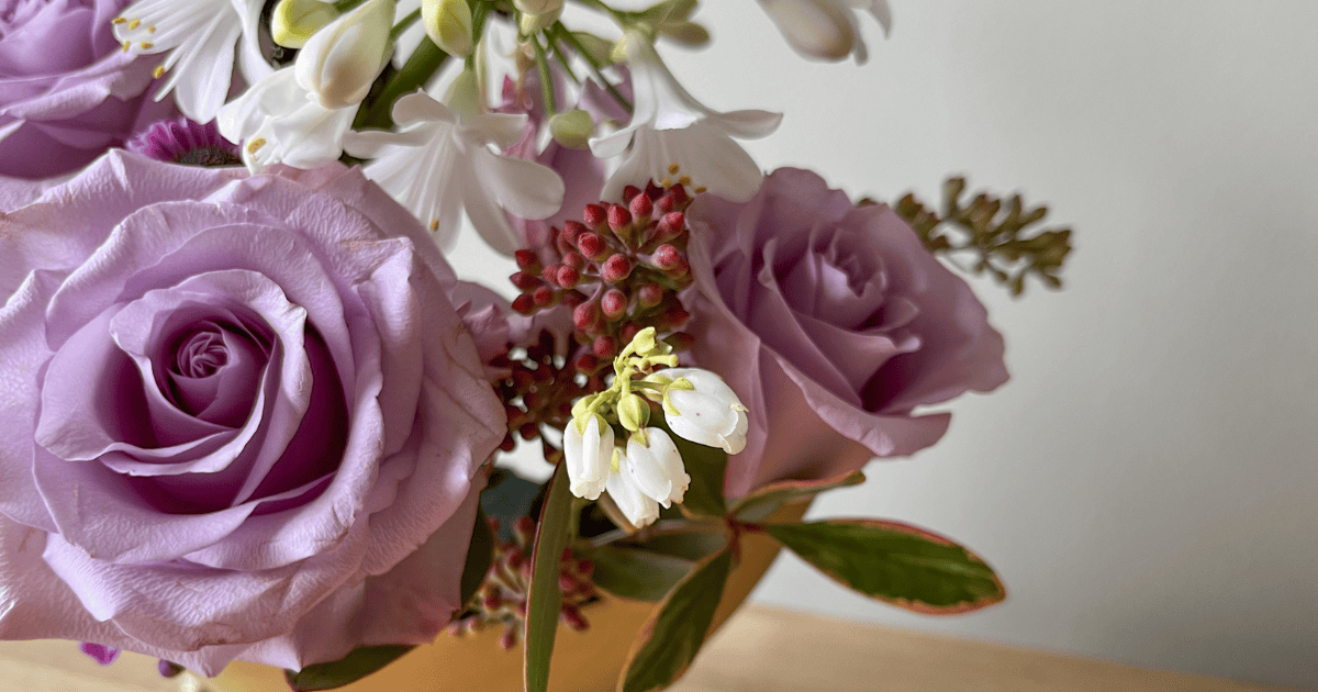 A close-up of a floral arrangement featuring lavender roses, white bell-shaped flowers, and clusters of pinkish-red buds on a light background.