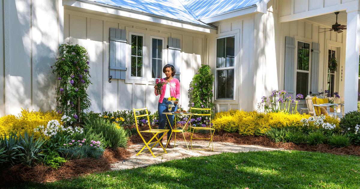 A person tends to flowers in a well-maintained garden with yellow chairs and a table, in front of a white house with blue shutters.