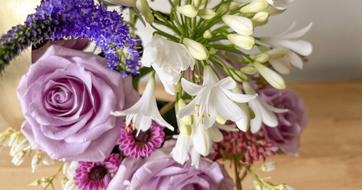 A bouquet featuring purple roses, white lilies, and other assorted flowers, arranged in a gold vase on a wooden surface.