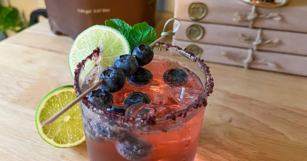 A pink cocktail garnished with blueberries, mint leaves, and lime slices is served in a glass with a sugared rim. It is placed on a wooden surface.