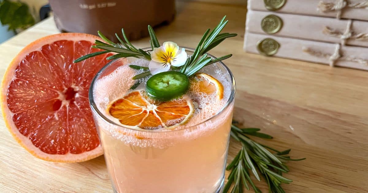 A cocktail garnished with slices of dried orange, a jalapeño, rosemary, and an edible flower, next to a halved grapefruit. Books are stacked in the background.