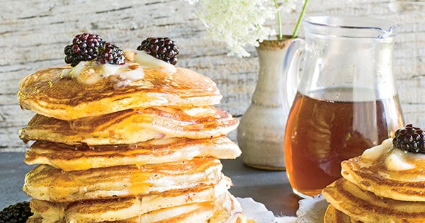 9 layer stack of pancakes topped with butter, syrup and blackberries and set on a table with pitcher of syrup and vase of flowers