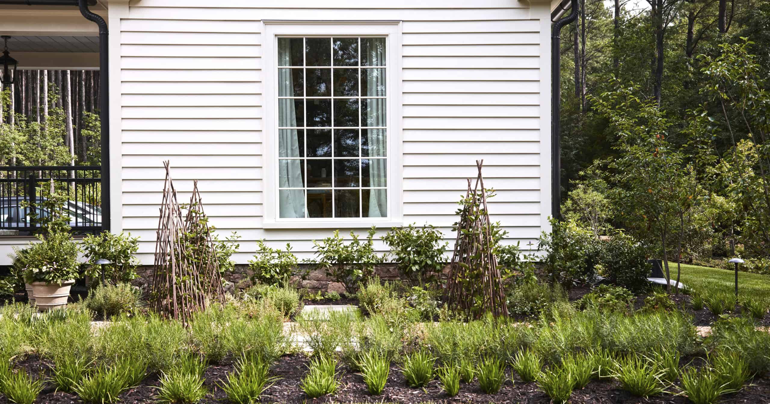 A vibrant kitchen herb garden along the side of the 2025 Southern Living Idea House. The space features neatly planted rows of Chef’s Choice® Culinary Rosemary, providing fragrant, evergreen texture. In the background, DownHome Harvest® ‘Osage’ Thornless Blackberry bushes grow along rustic wooden trellises flanking a central window. Additional herbs and edible plantings fill the beds, while the surrounding orchard trees and soft lawn extend the productive garden space into the broader landscape.