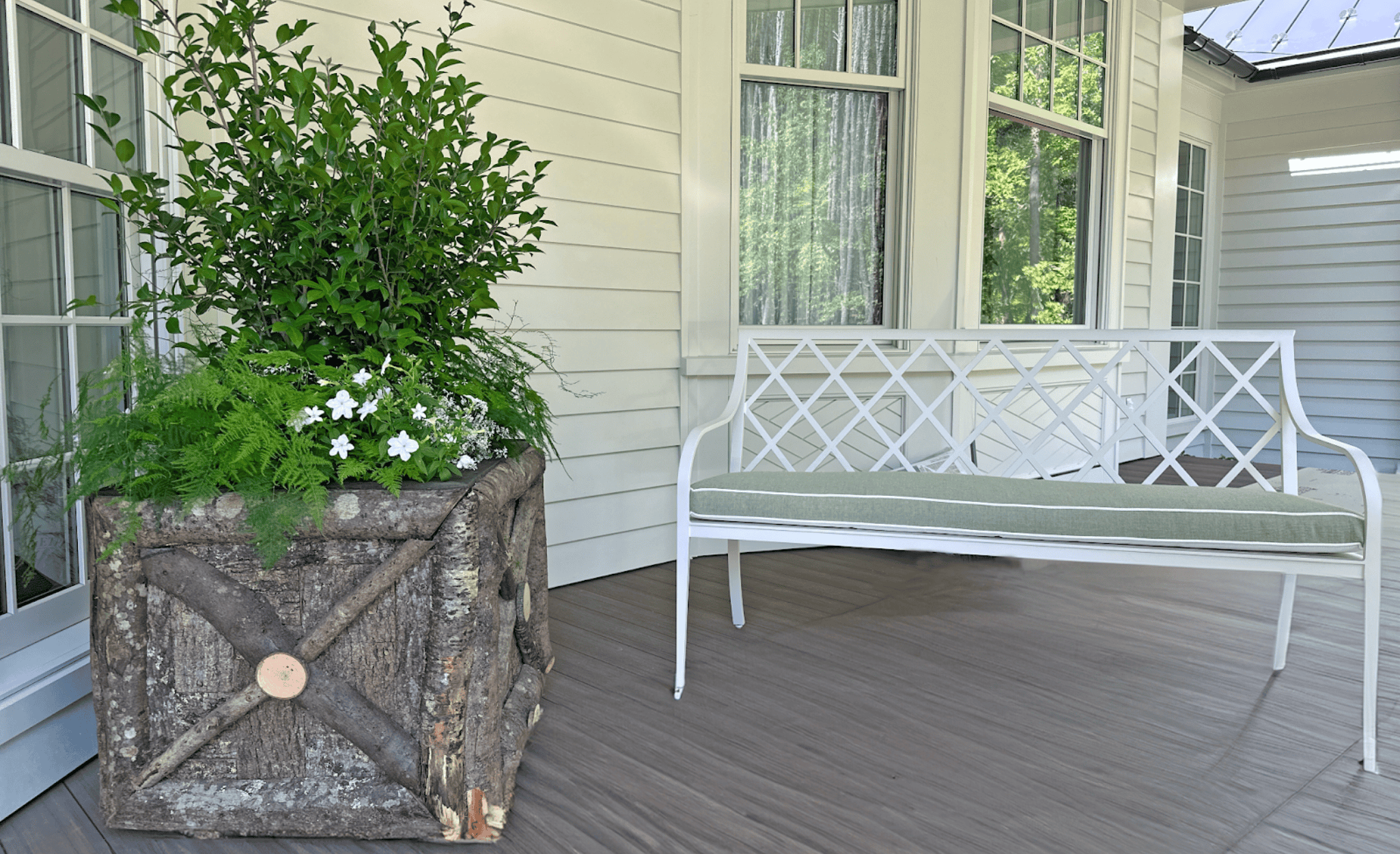 Rustic wooden log planter with October Magic® Ivory™ Camellia shrub, lush ferns, and white-blooming euphorbia beside a white metal bench with green cushion on the wraparound porch of a white house.