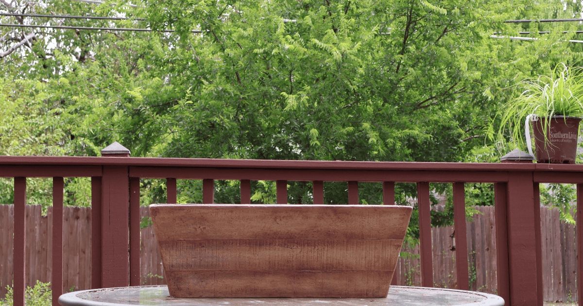 A wooden planter sits on a glass patio table with a wooden fence and green trees in the background.